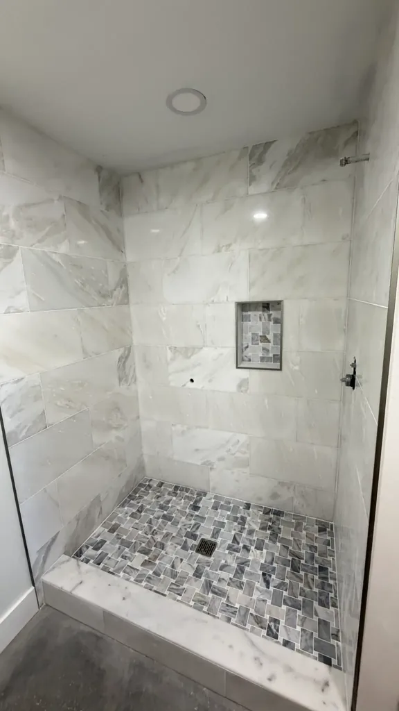 Angled view of shower wall tile layout, mosaic floor, center drain and light veined curb