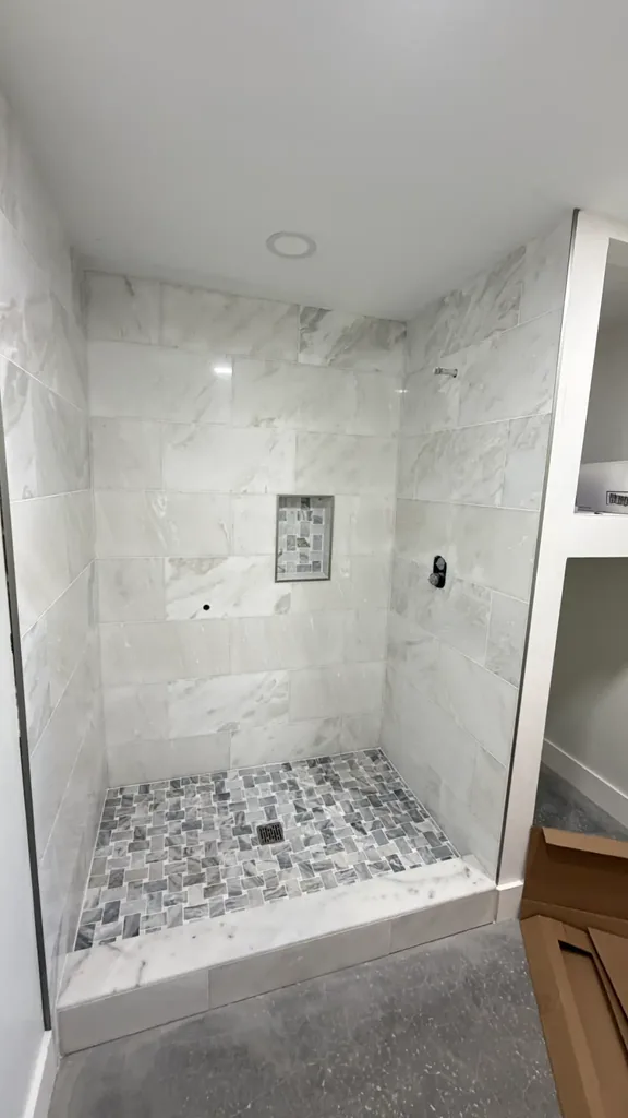 Wide view of installed light veined shower wall tile, gray mosaic floor, recessed niche and tiled curb