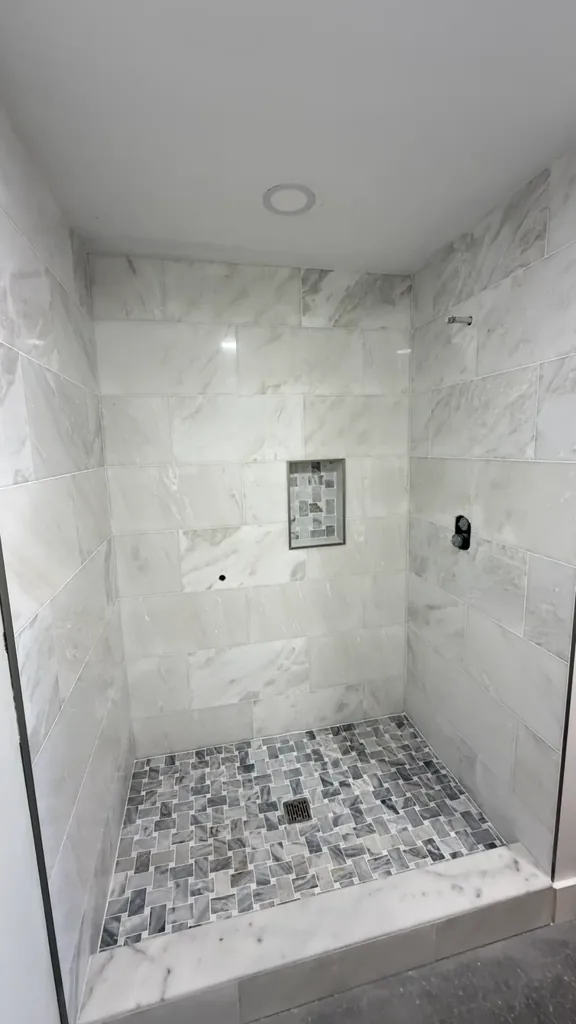 Front view of the tiled shower with light rectangular wall tiles, contrasting mosaic floor and recessed niche