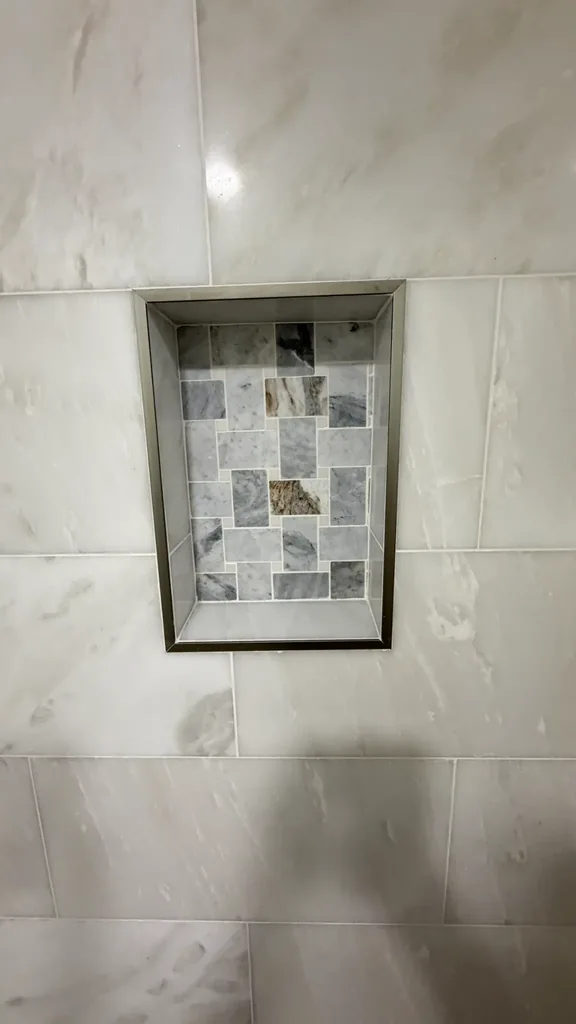 Close view of recessed shower niche with gray and white mosaic backing and contrasting border trim